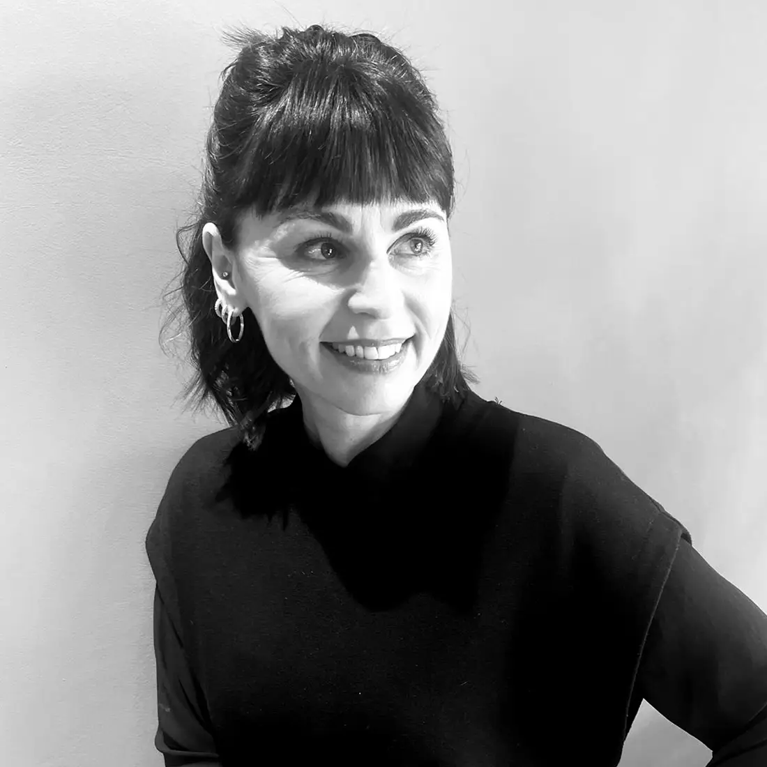 Portrait von Carolin in Schwarz-Weiß, lächelnd mit Pony und zurückgebundenem Haar, Blick zur Seite, Ohrringe, neutraler Hintergrund.