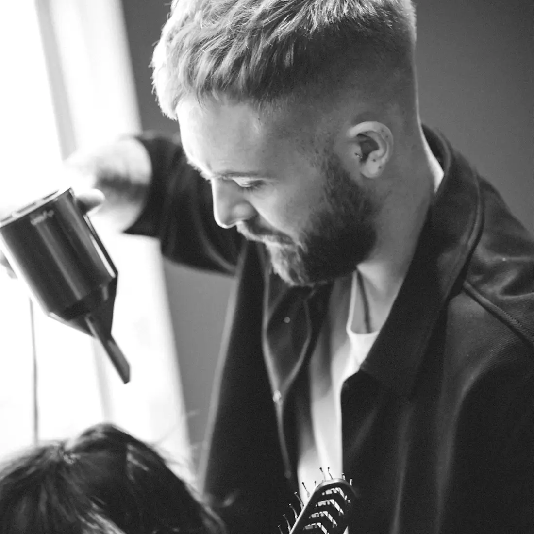Marco Stauber föhnt das Haar einer Person; Nahaufnahme in Schwarz-Weiß mit Föhn und Stylingkamm im Vordergrund.