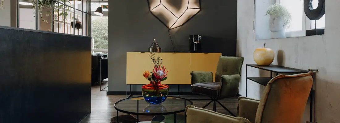 Moderner Wartebereich im Salon Marco Stauber mit samtigen Sesseln, rundem Glastisch und farbiger Vase; gelbes Sideboard und Designleuchte im Hintergrund, rechts eine raumhohe Abtrennung.